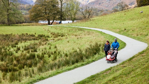 A young family walk with a pushchair on a tarmac path snaking down from the fells at Langdales, Cumbria. The wintery hills are behind them, along with a lake boarded by bare trees.